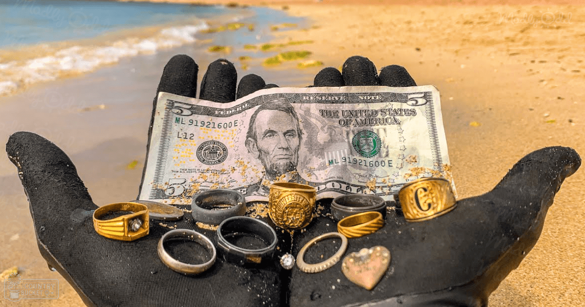 Hand in rubber gloves holding jewelry and a five-dollar bill on a beach. Daytime scene with sand, water, black gloves, gold rings, silver pendant, beige sand.
