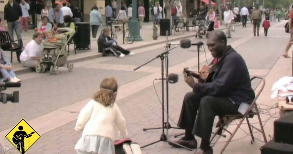 An adult male street musician in a dark suit jacket and white shirt plays an acoustic guitar on a sunny urban plaza with various pedestrians and strollers, surrounded by neutral tones and pops of color.