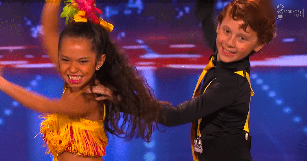 A young female dancer wearing a yellow fringed top and skirt with a floral headpiece stands beside another child, dressed in a black and yellow dance costume on a stage with blue and red backdrops.