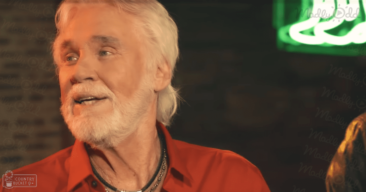 An older adult male wearing a red button-up shirt and black chain necklace, set against an indoor backdrop with dim lighting and a partially visible neon sign.