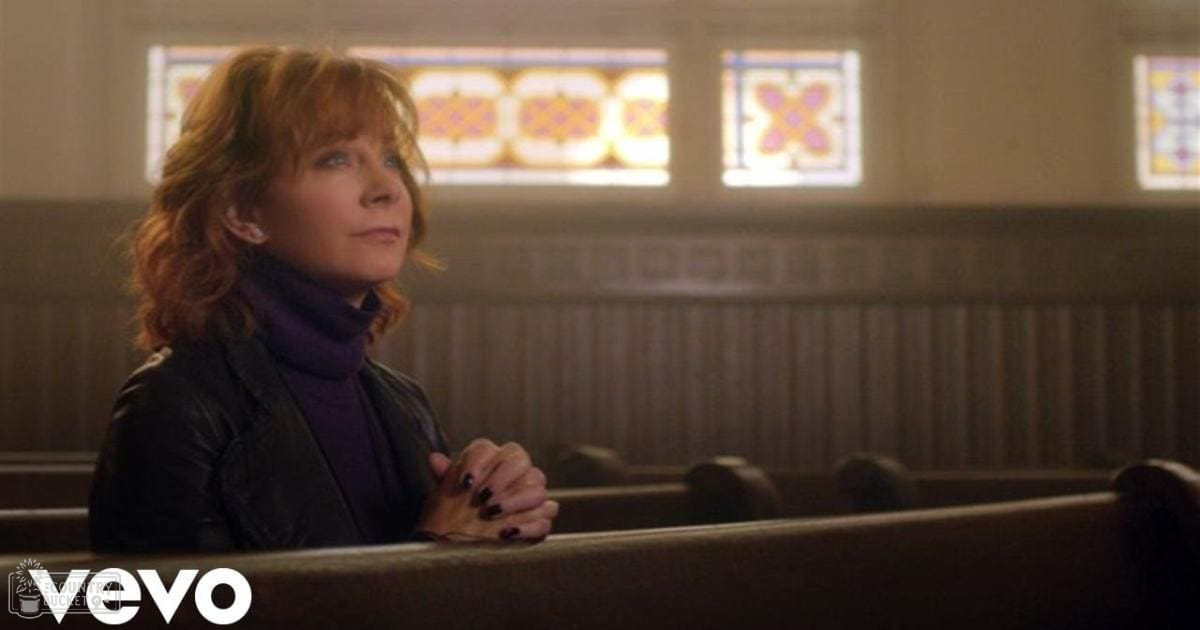 A middle-aged woman wearing a black leather jacket with high collar and turtleneck in a church setting, surrounded by wooden pews and stained glass windows. The scene suggests a peaceful daytime atmosphere.