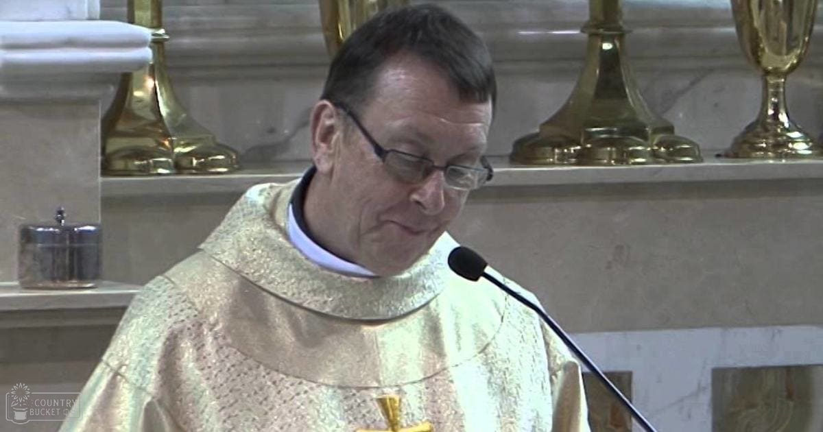 A middle-aged adult male in a church setting, wearing a priest's alb with subtle embroidery and holding a small object.