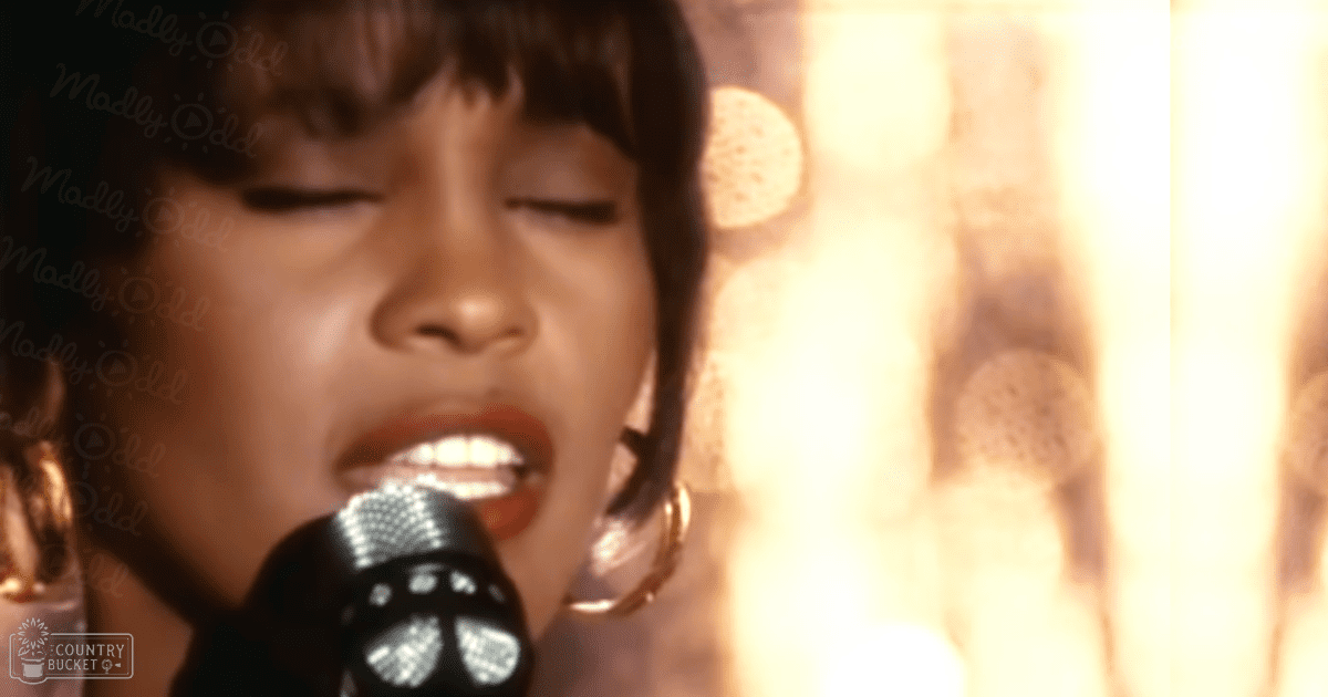 A young adult female singing into a microphone indoors with stage lighting in warm tones.
