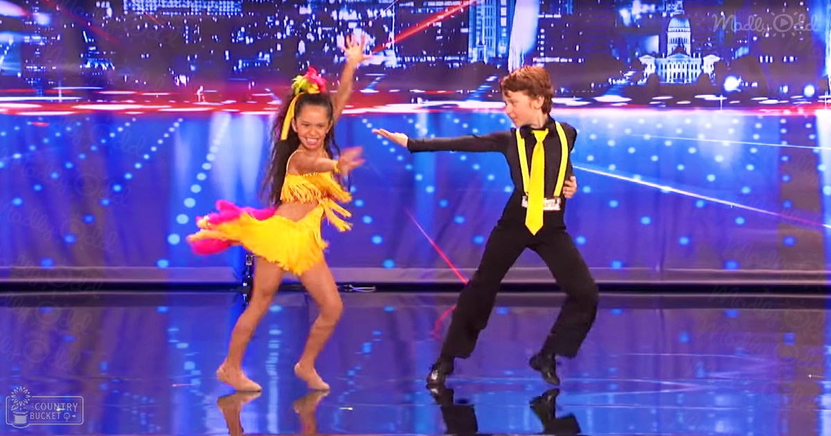 A young couple dancing on a glossy stage with a cityscape backdrop at night, wearing bright yellow fringed dresses and black formal attire under artificial lighting.
