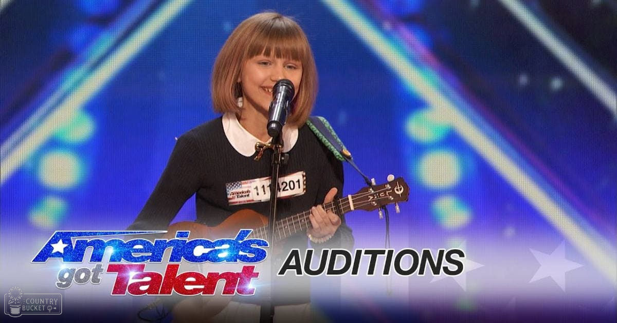 A young girl wearing a black sweater with white collar performs at an 'America's Got Talent' audition in a studio setting, holding an acoustic guitar and standing before a microphone stand.