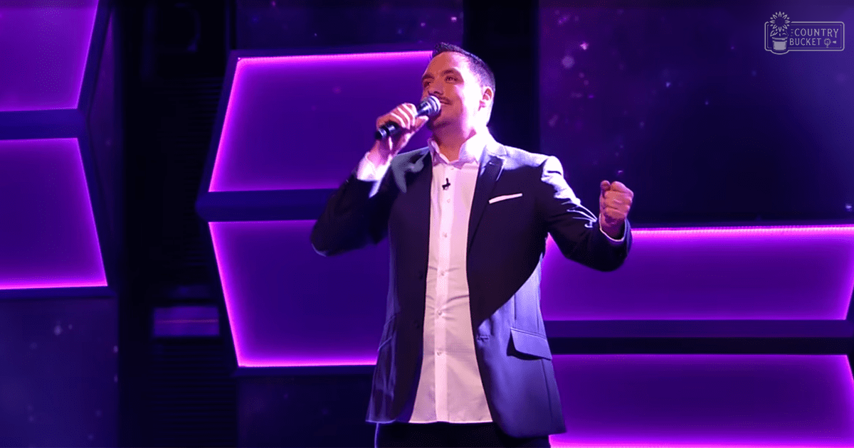 An adult male performer in a dark blazer and light button-up shirt on an indoor stage with purple lighting.
