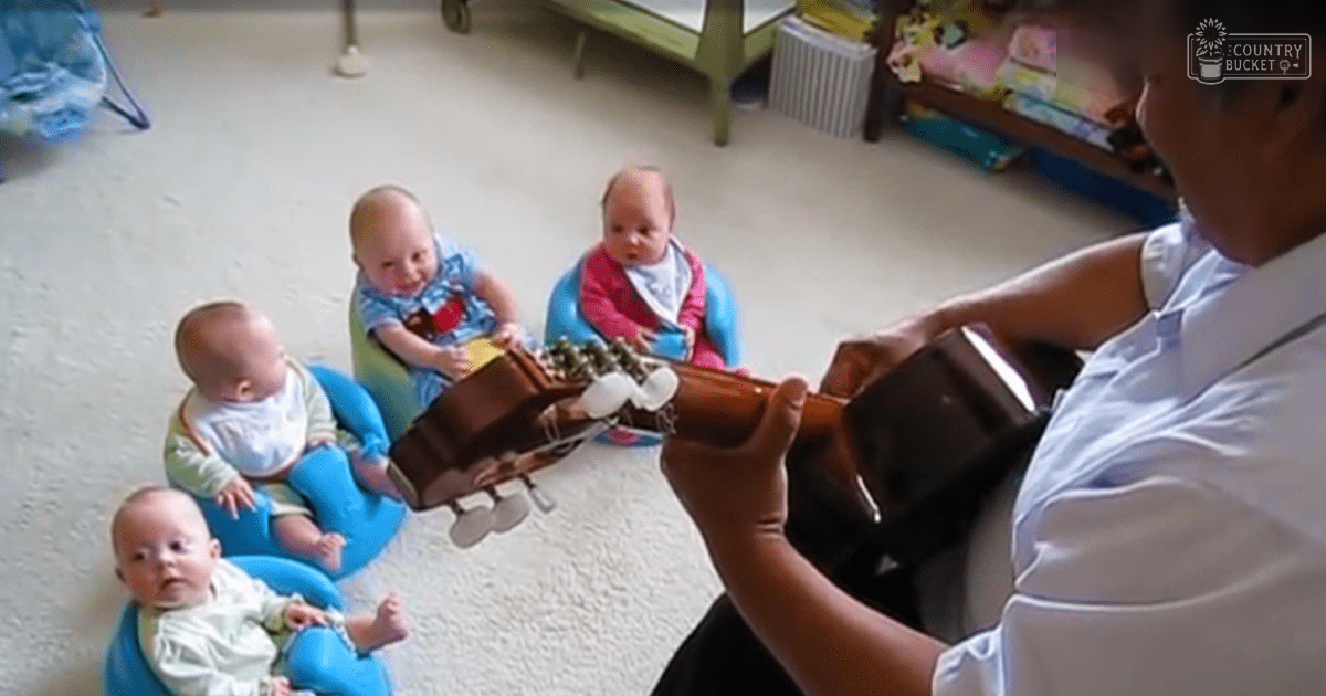 Adult male playing guitar for infants indoors, light blue shirt, green table with white radiator, natural daylight.