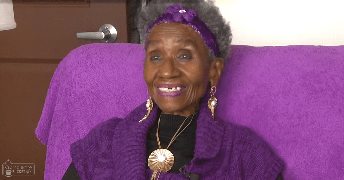 An older adult woman wearing a purple textured sweater and large hoop earrings sits indoors with a purple cushioned chair.