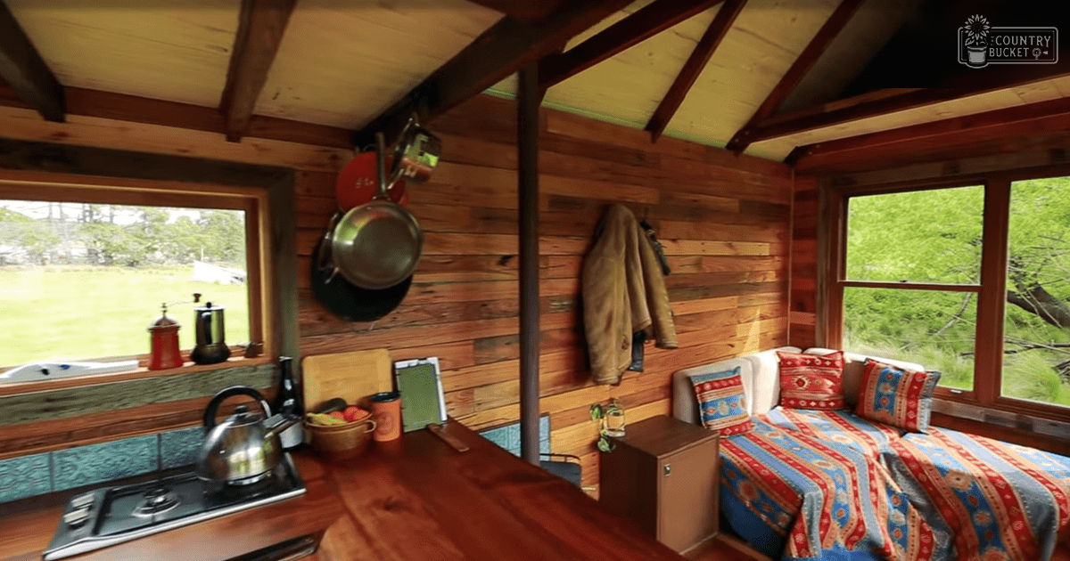 Cozy cabin interior with wooden walls, colorful bedding, kitchenette, and lush greenery outside.
