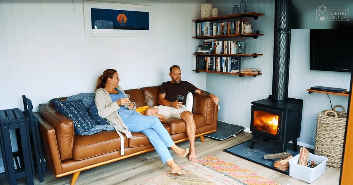 A middle-aged couple relaxing in a cozy living room with blue floral cushions, a glowing wood-burning stove, and natural daylight illuminating spring or summer attire.