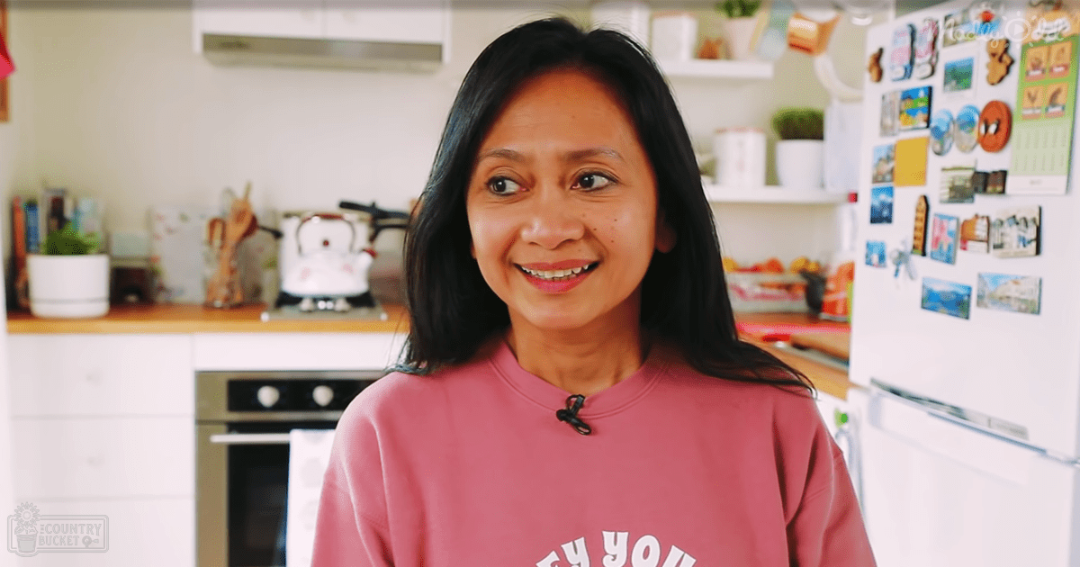 A middle-aged woman in a modern casual pink sweatshirt stands amidst kitchen furnishings, surrounded by natural light.