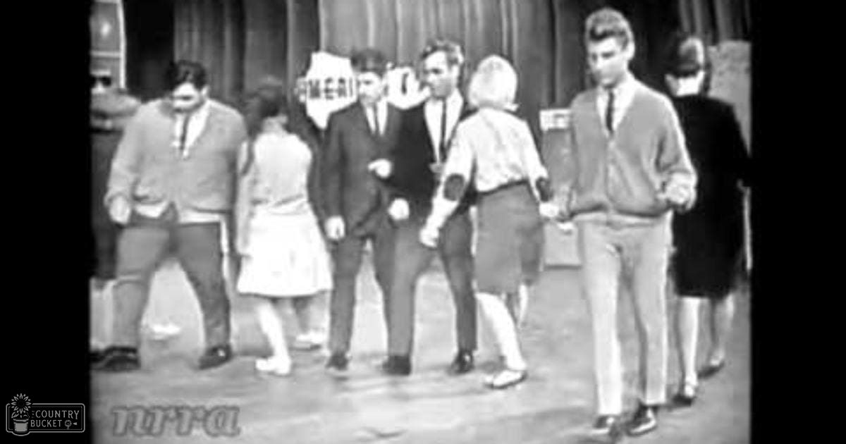 Adult male wearing light sweater with dark patterned tie, child in white dress or skirt, adult males in suits and casual attire on stage set for performance.