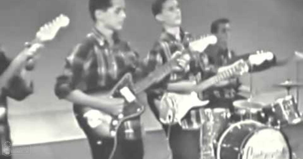 A musical performance with young adult males playing guitars and drums indoors, likely at a music venue or school auditorium, with muted lighting and monochromatic colors.