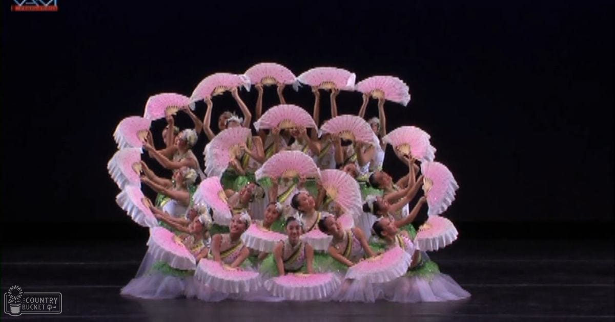 A group of young adult female dancers in green and white costumes with ruffled skirts perform on a dark stage under artificial lighting.