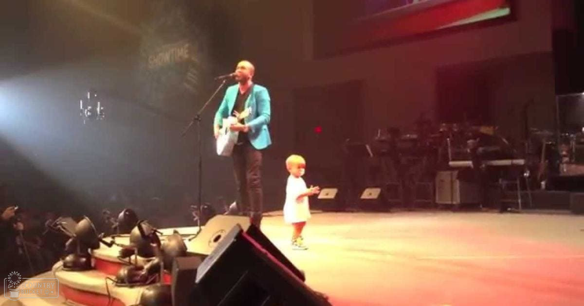 A young child in a white dress stands on stage with an adult male wearing a teal blazer and dark pants during what appears to be a concert performance at night.