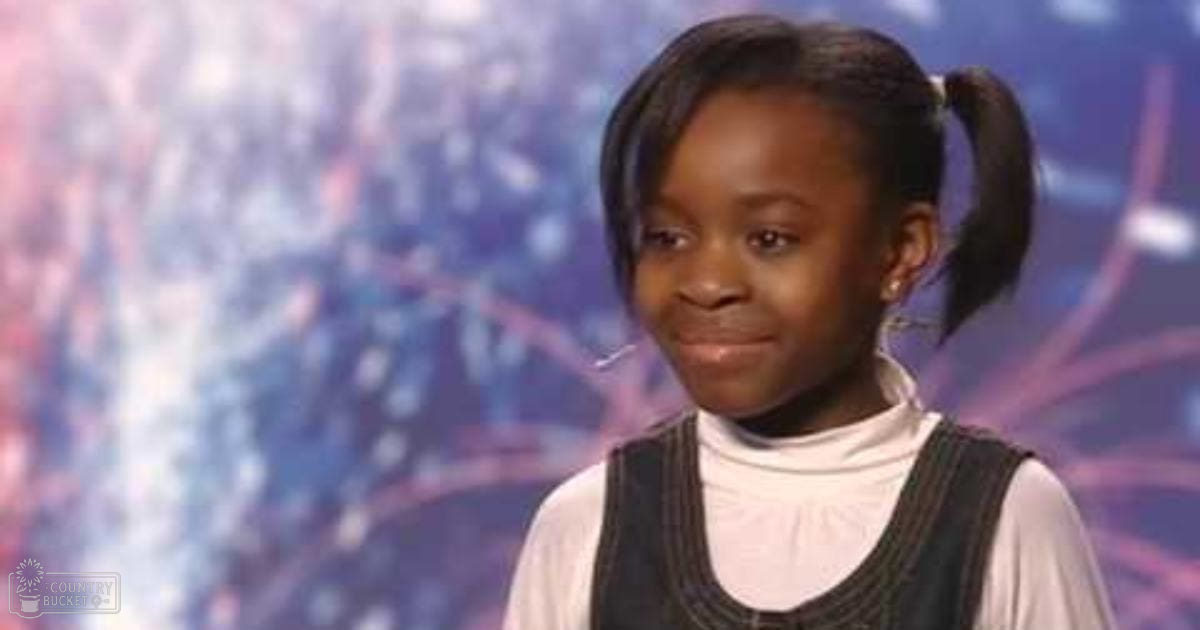 A young child with dark pigtails in a white turtleneck sweater and black vest, standing indoors at an evening performance venue.