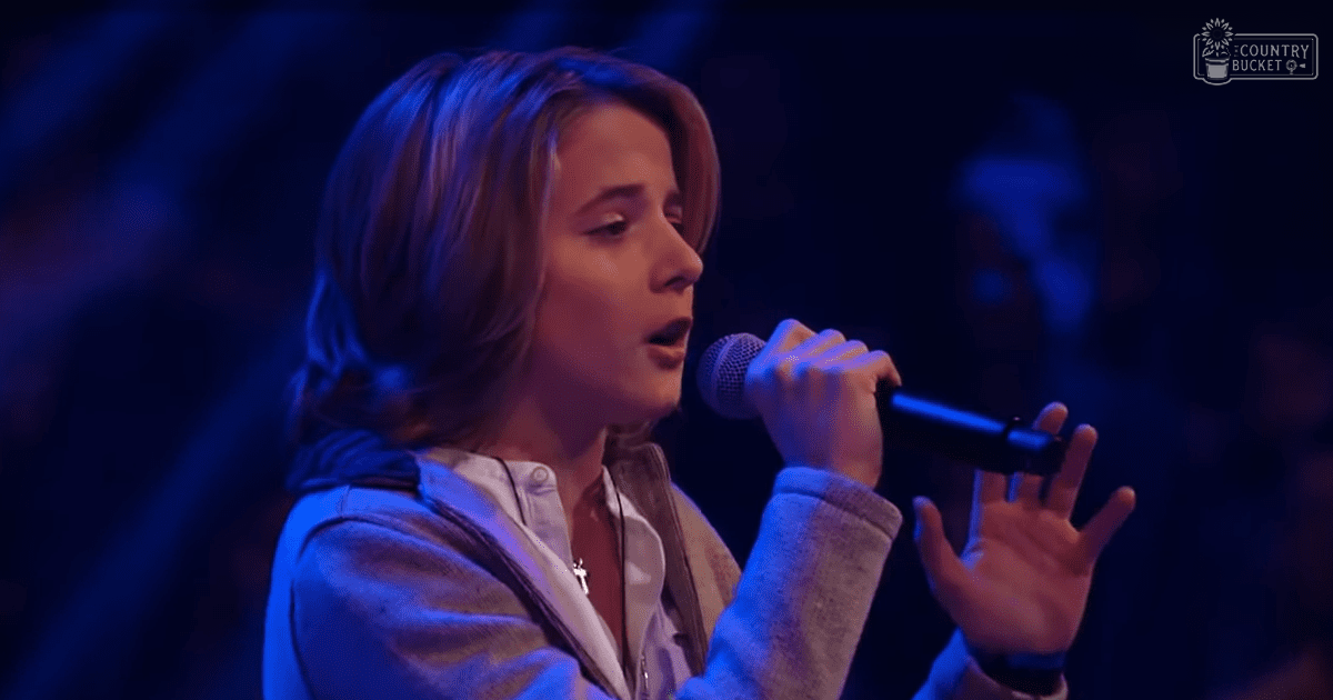 A young adult female sings into a microphone at an indoor performance event, wearing a light-colored shirt with a gray zip-up hoodie layered over it and holding a cross necklace.