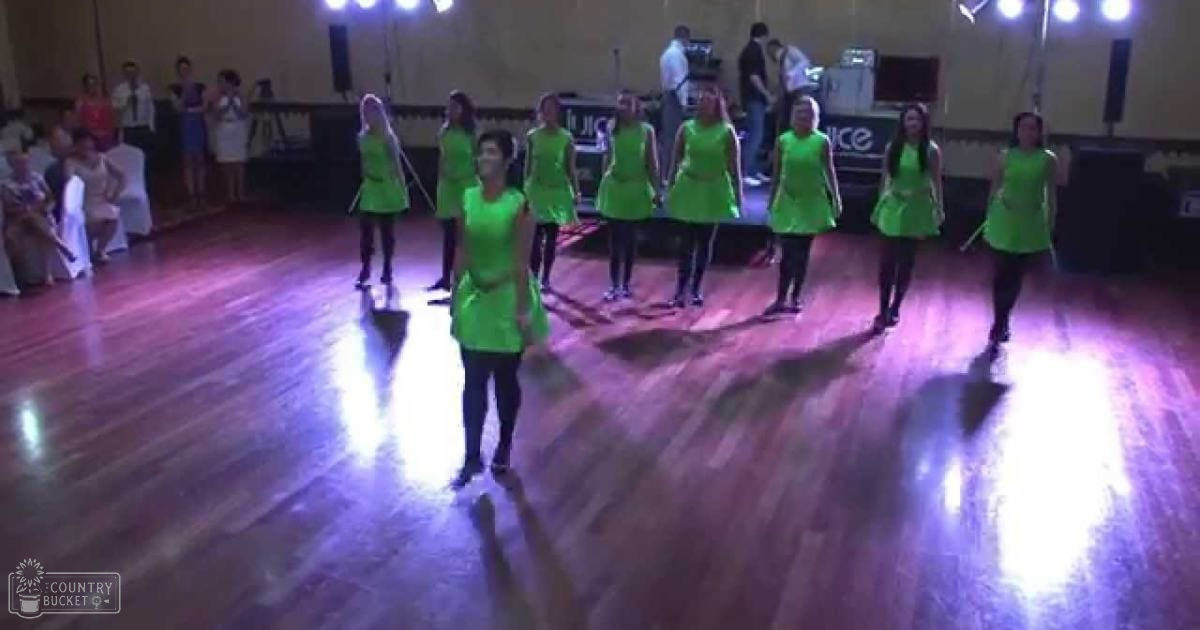 Young adult females in sleeveless knee-length green dresses performing at an indoor dance venue with wooden flooring and stage equipment visible.