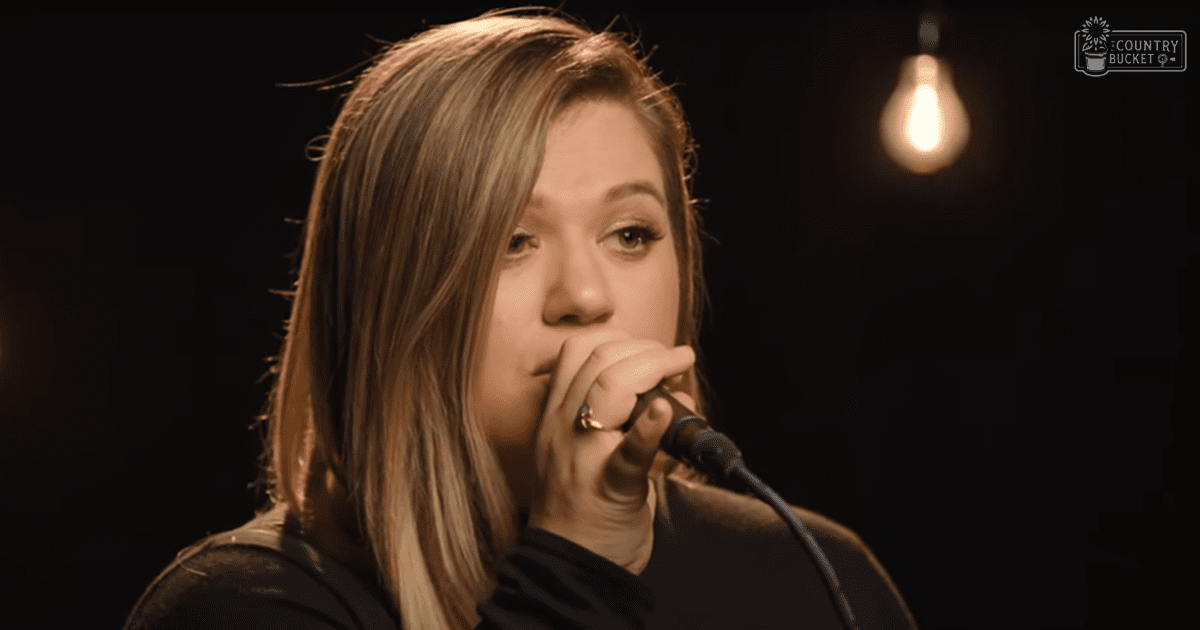 A young adult female in a long-sleeved black top, singing into a microphone indoors under warm lighting.