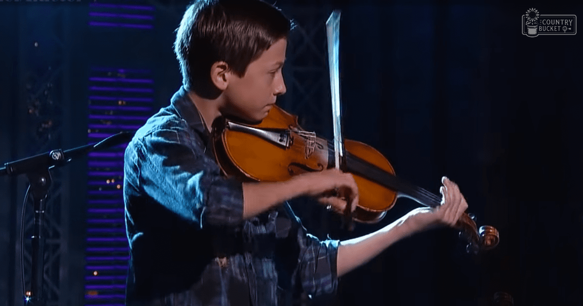 A young boy in a blue plaid shirt plays the violin on stage under purple lighting, wearing regular fit, slightly loose long-sleeved button-down shirt made of cotton or similar lightweight fabric.