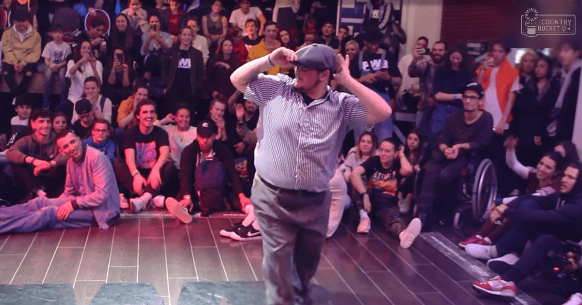 A middle-aged male dancer in a striped button-up shirt and khaki pants performs on stage during an indoor dance event, with an audience of young adults seated around him.