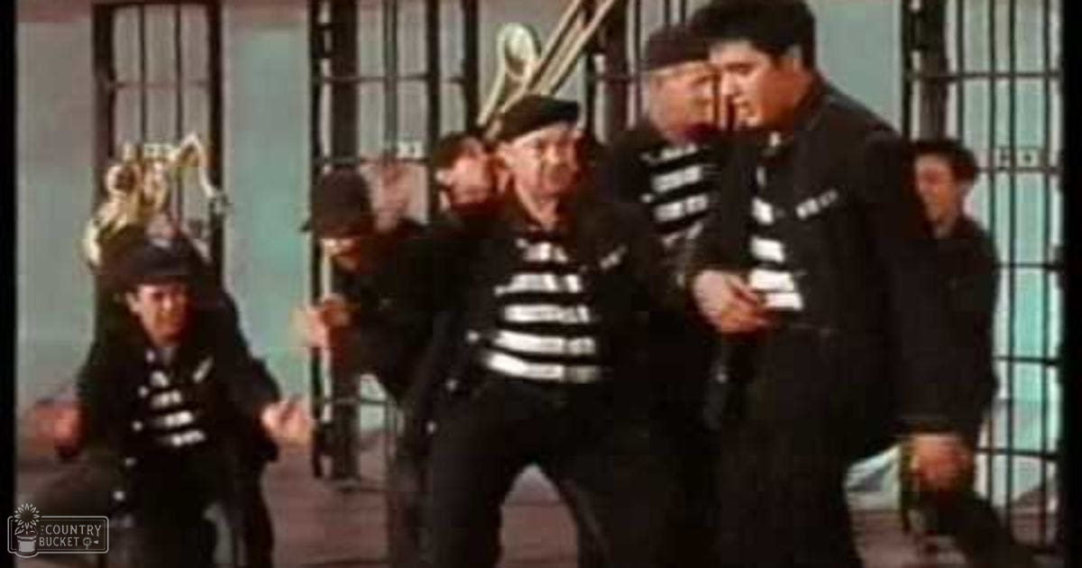 A group of adult males in prison uniforms stands outside a jail cell, dressed in striped shirts and dark pants, with some holding weapons. The setting suggests an urban environment typical of mid-20th century films.
