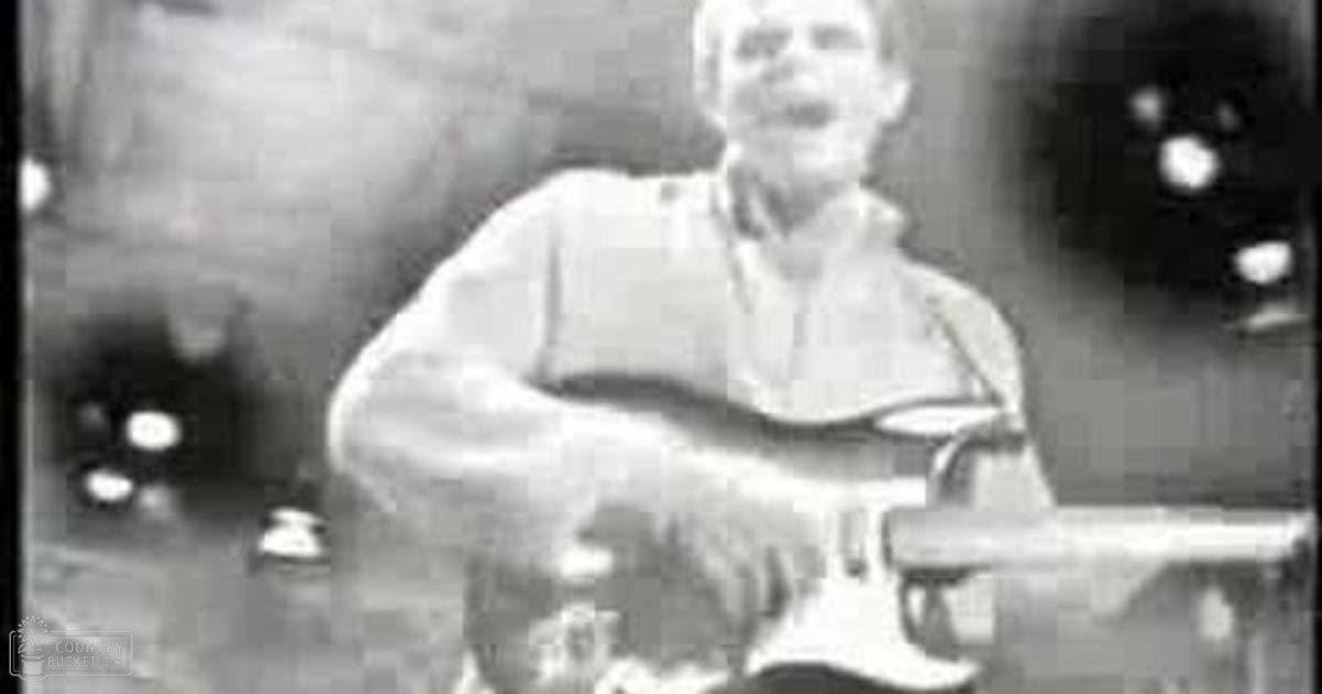 A middle-aged adult male plays an electric guitar in a dimly lit bar or music venue.