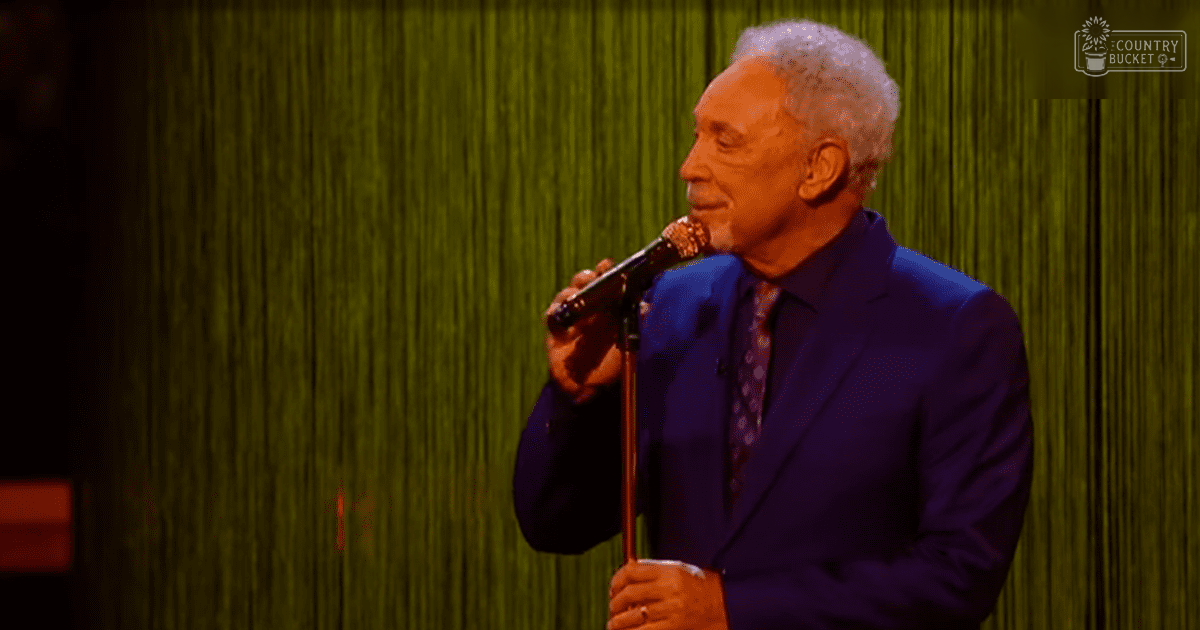 An older adult male in a dark purple suit holding a microphone, performing on stage with green curtains and 'Madly Odd' logo visible.
