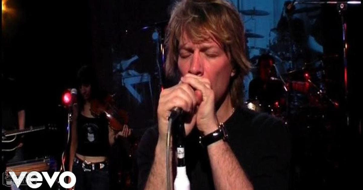An adult male singer wearing a black short-sleeved shirt, wristwatch, and bracelet sings into a microphone on stage at a concert with other musicians in the background.