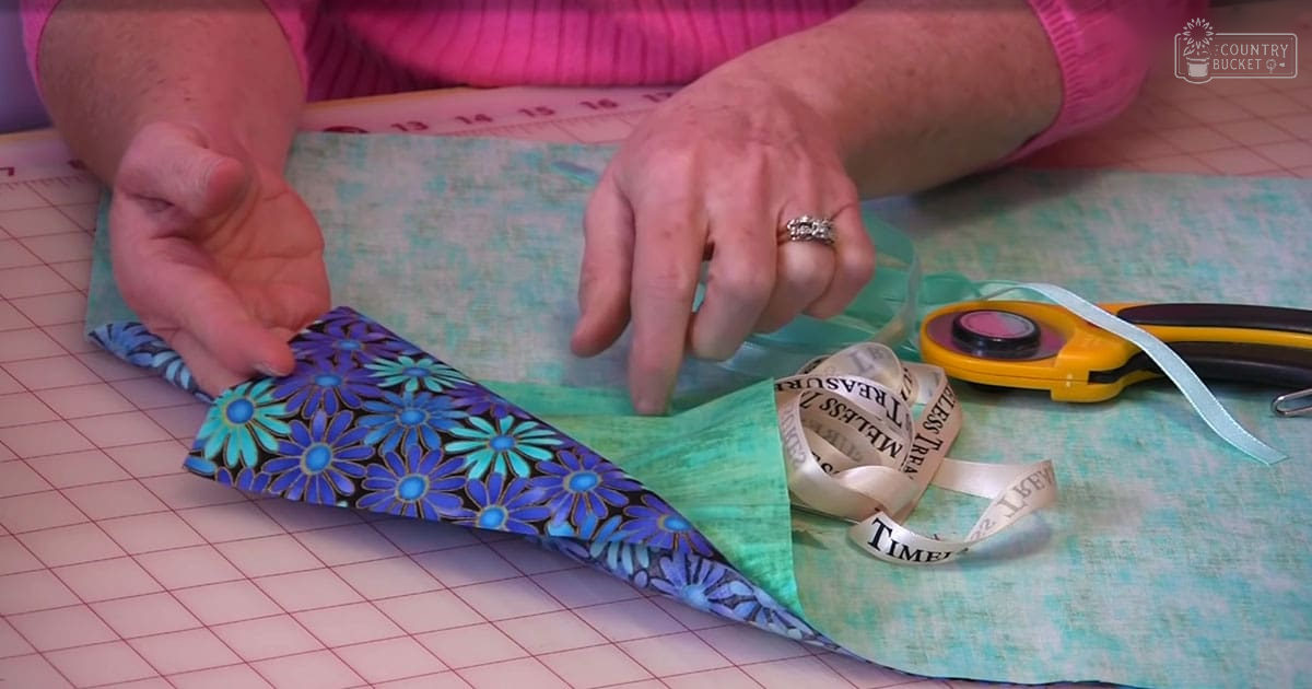 A middle-aged adult female engaged in crafting at home, surrounded by tools like a rotary cutter and fabric pieces with various patterns.