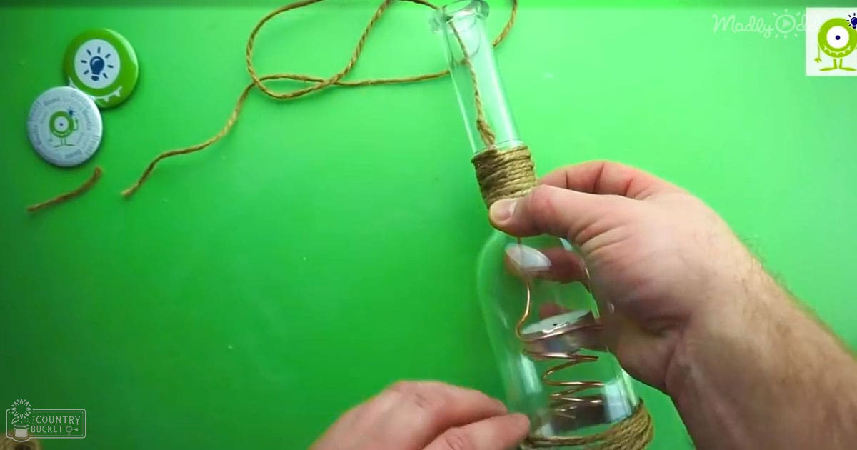 Adult male crafting twine around glass bottle against green backdrop, two branded buttons visible on workspace.