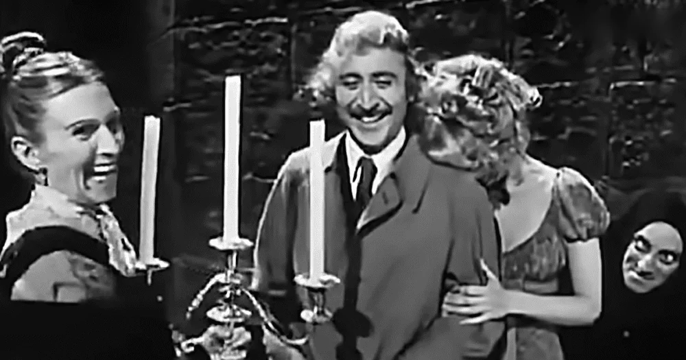 Adult male wearing dark suit jacket indoors with young adult female on left and another adult female partially obscured, possibly part of a theatrical scene.