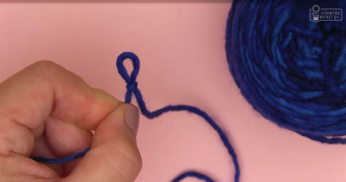 A hand holding blue yarn against a plain pink surface, with 'Molly Odd' text visible in the upper right corner.