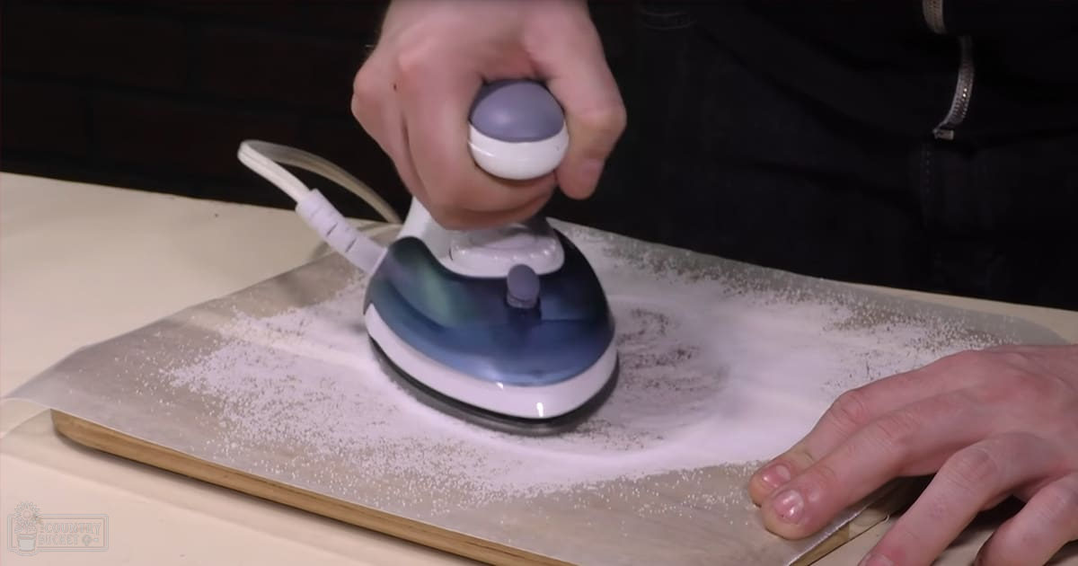 An adult male wearing a dark-colored jacket or sweater is ironing fabric on a wooden cutting board in an indoor setting with artificial lighting.