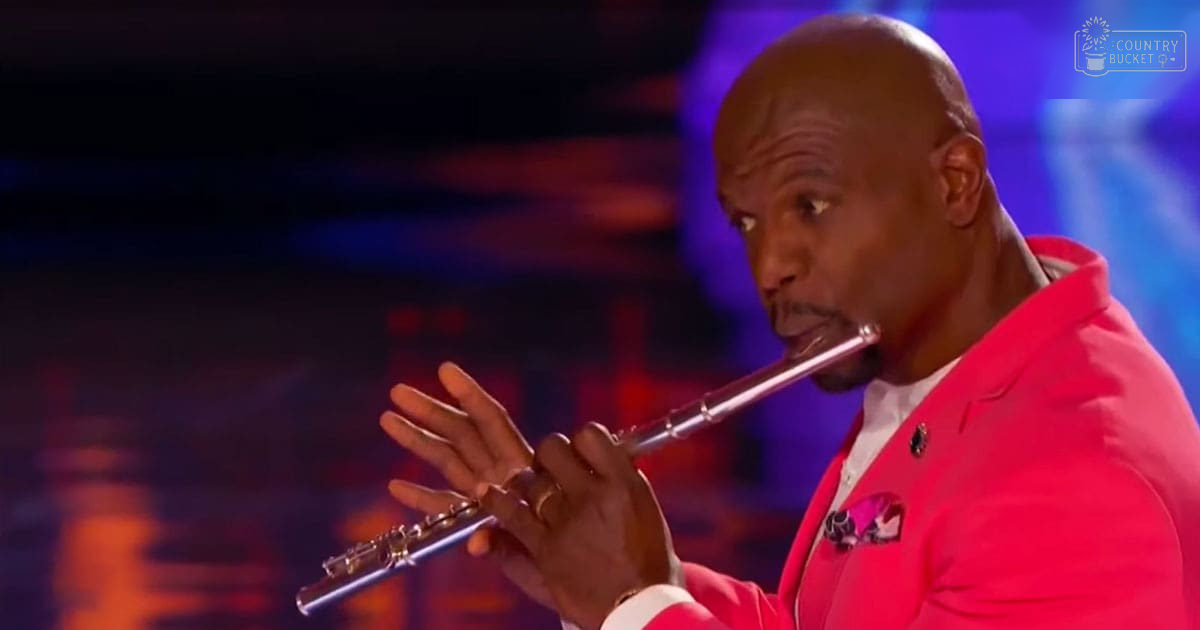 An adult male singer wearing a bright pink blazer with a white shirt, playing a metallic silver flute on stage at an event.
