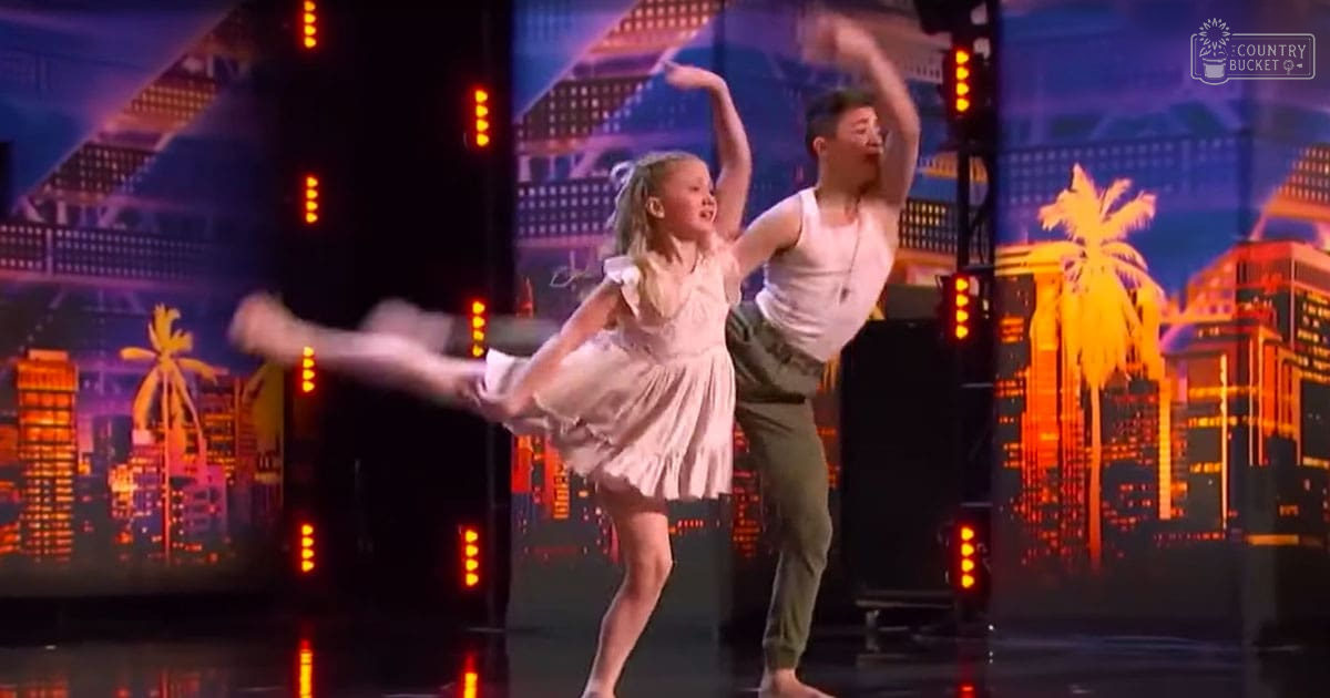 A young girl performing ballet with an adult male dancer on stage against a vibrant cityscape backdrop; evening lighting enhances the colorful performance setting.