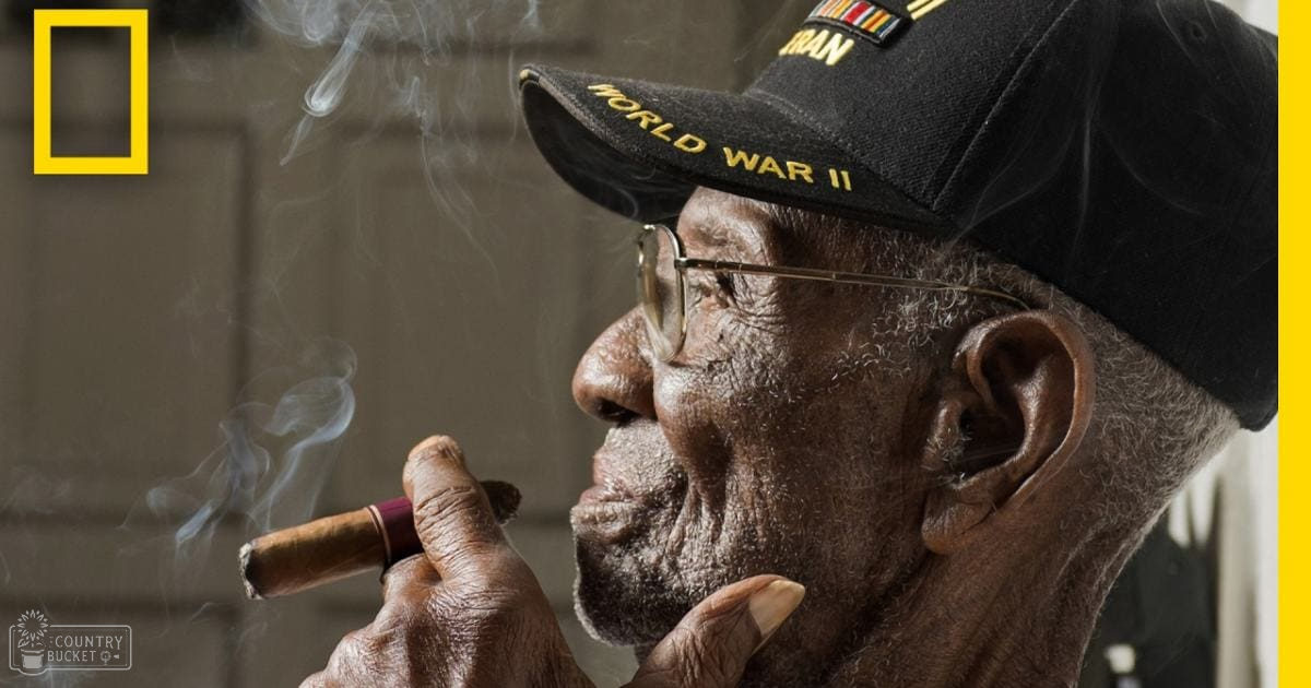 An older adult male wearing a navy-style cap with 'World War II' and 'Iran' patches, smoking a cigar indoors under natural light.