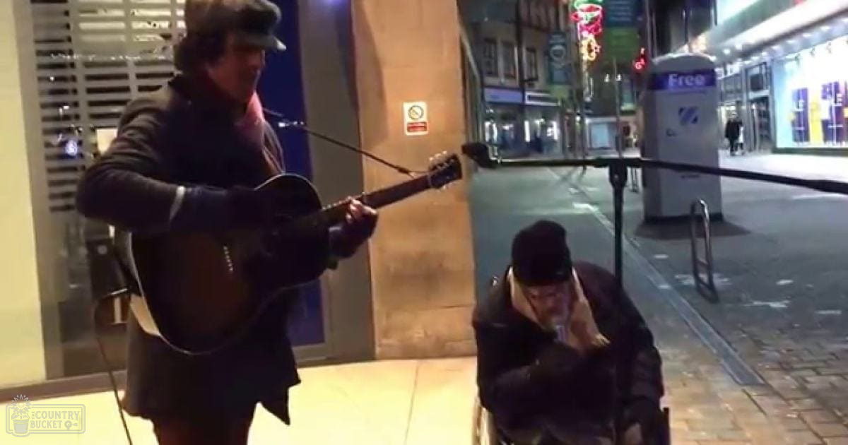 A street musician plays guitar while another person sits nearby under artificial lights at night on an urban street, surrounded by illuminated signs and a brick-paved ground.