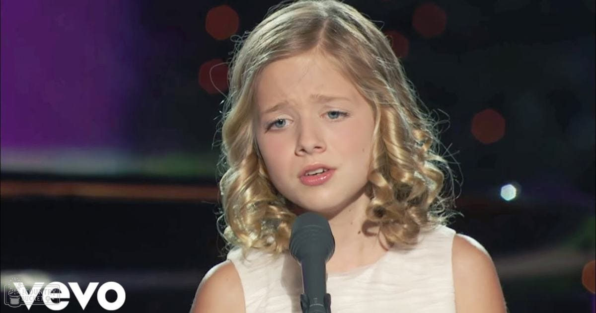A young female child wearing a white sleeveless top, holding a microphone in an indoor setting with stage lighting.