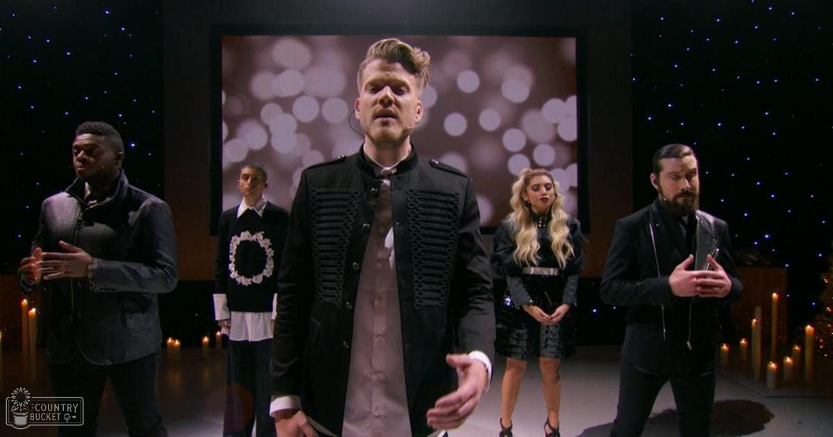 An adult male singer in a black jacket with decorative silver buttons, surrounded by other young adults at an indoor performance venue with starry effects and bokeh lights.