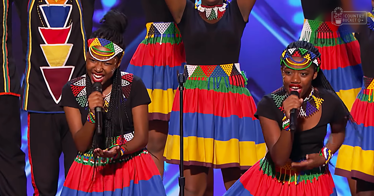 Two young adult female performers in traditional African attire with intricate beadwork and vibrant colors singing on a brightly lit stage.