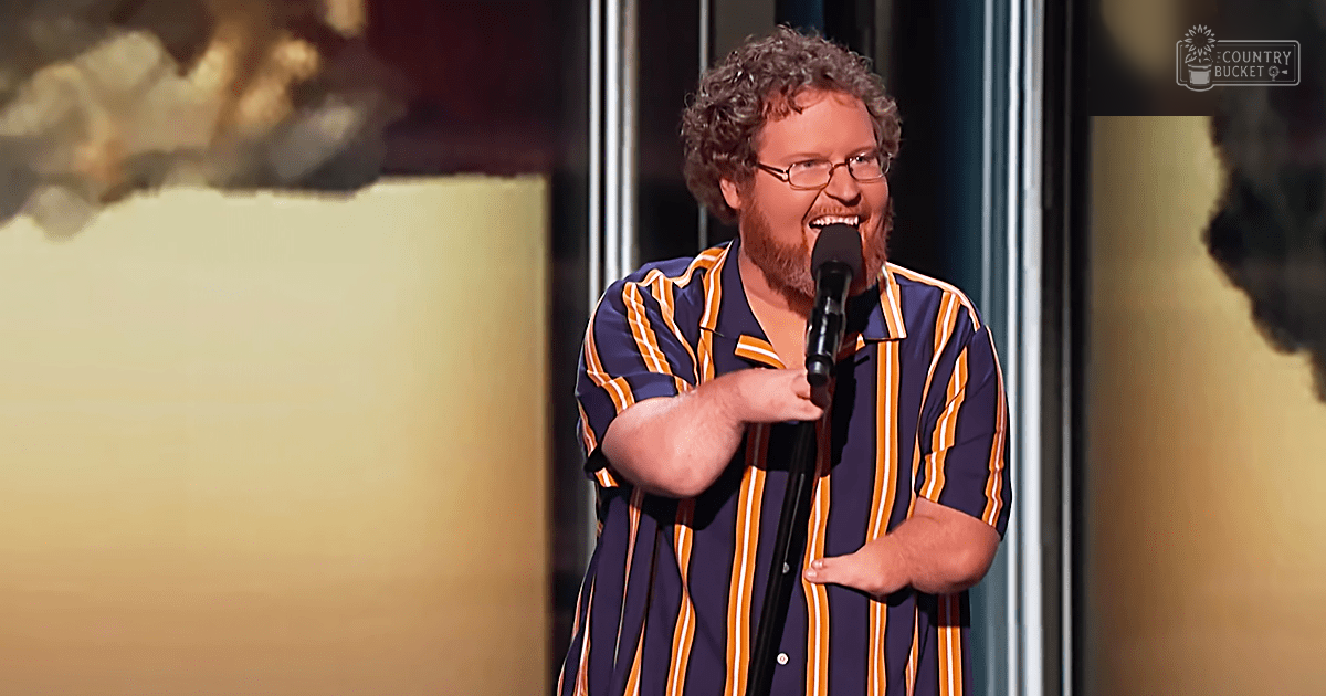 A male comedian performs stand-up comedy indoors, wearing a short-sleeved striped shirt with blue and orange vertical stripes. A microphone stand is prominent in the foreground.