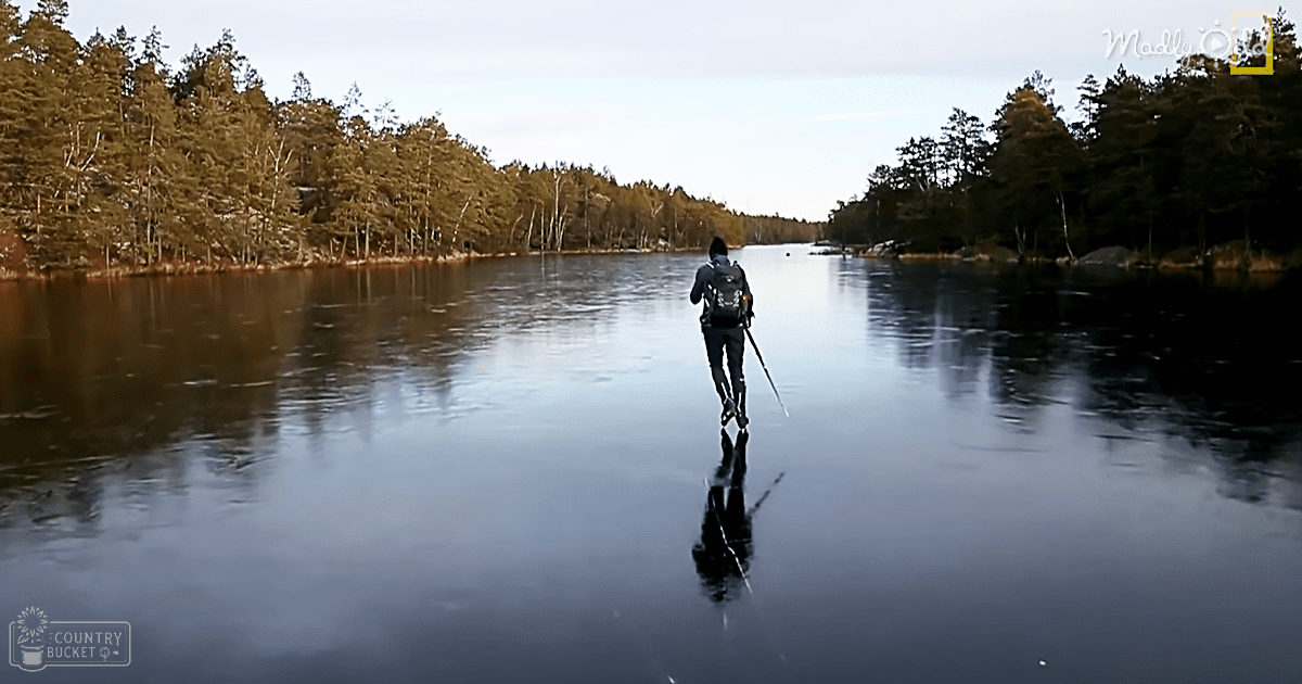 An adult male walking across a thin, icy lake surrounded by dense forest under an overcast sky during early spring.
