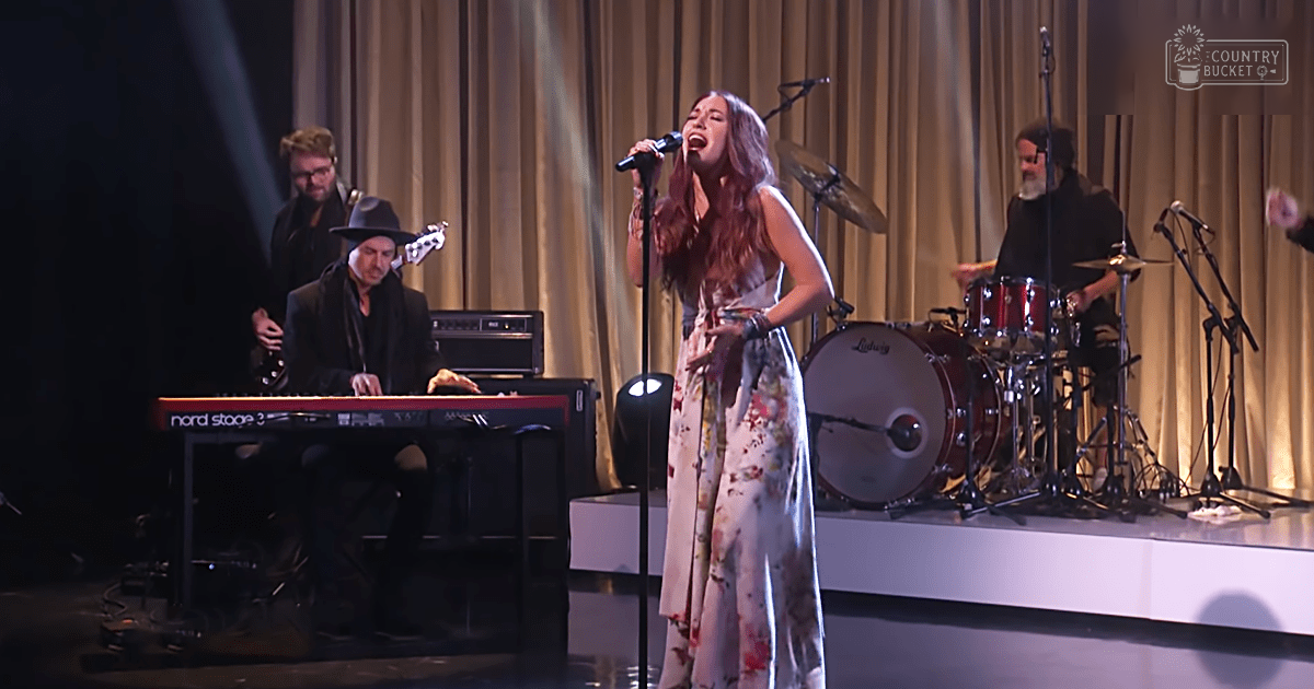 A young adult female singer in a floral dress performs on stage at an indoor venue with other musicians and stage equipment visible.
