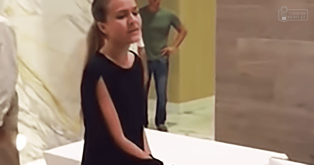 Young adult female wearing dark blue sleeveless dress with round neckline and fitted cut stands indoors; adult male in green t-shirt and jeans behind her, hands on hips.