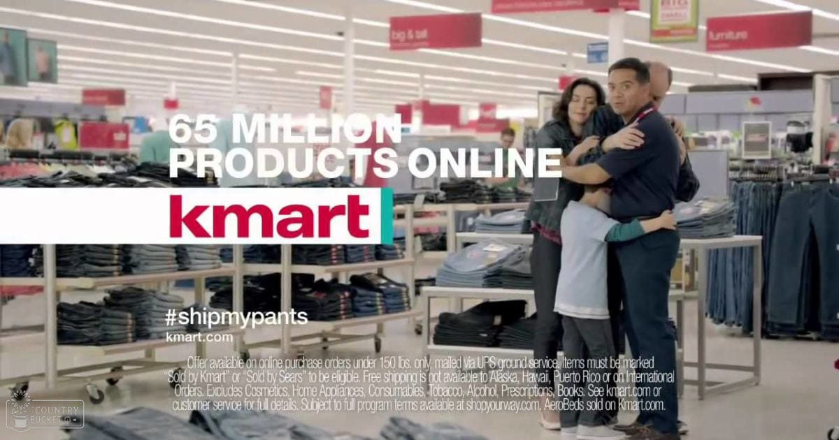 An adult male singer in a dark-colored short-sleeved shirt and dark pants embraces an adult female wearing a light-colored top with long sleeves, both surrounded by shelves of clothing items in a Kmart store.