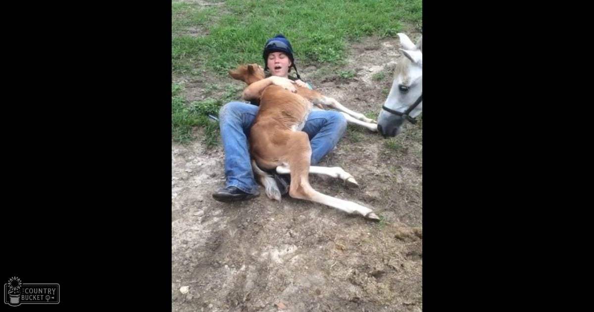 A young adult female in a blue jeans and black riding boots, wearing a backward blue helmet or cap, stands outdoors on grass with animals: a brown and white dog lying down and a white horse standing nearby. The setting appears to be spring or summer.