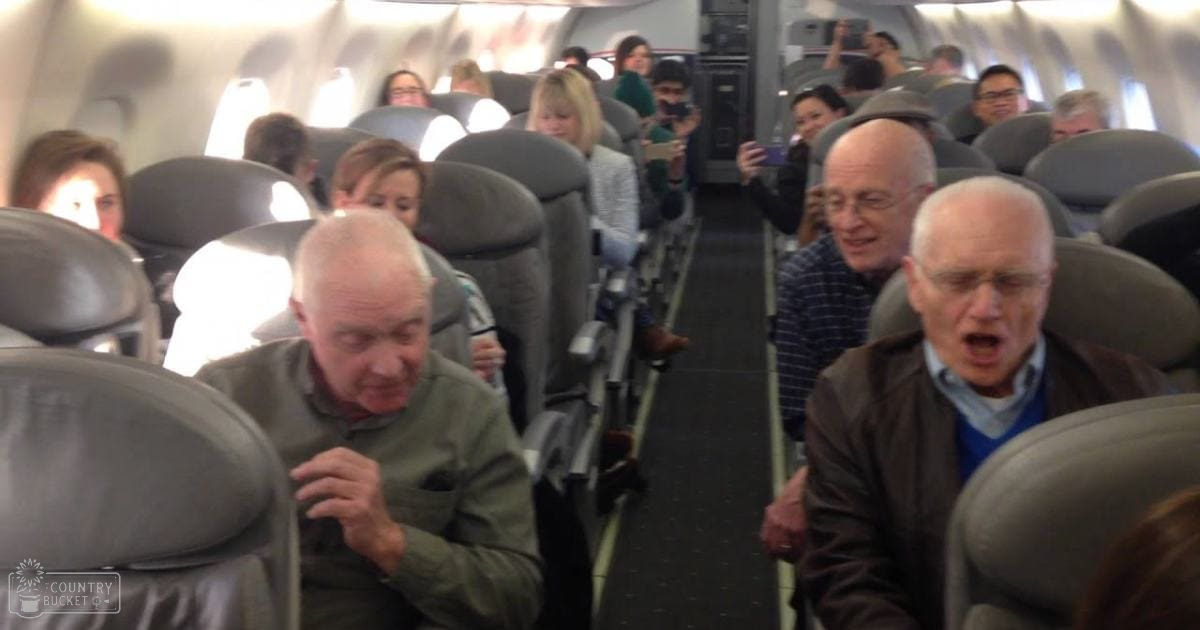 Middle-aged man in green jacket seated inside an airplane cabin during a flight with natural daylight streaming through the windows.