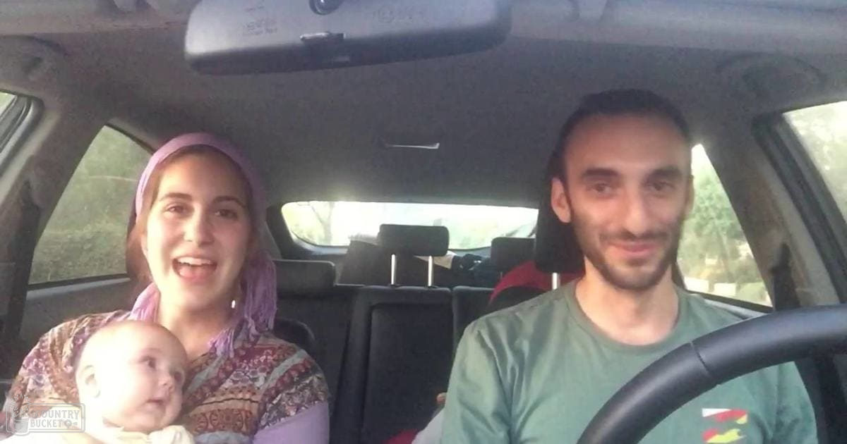 A family inside a car, with an adult female holding a baby and wearing a patterned top with a purple headscarf; the driver is an adult male wearing a green T-shirt. Trees outside suggest they are on a road trip during daylight hours.