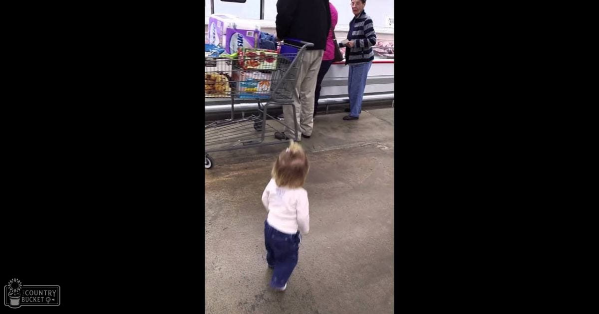 A toddler in a white shirt with text on it stands near an adult male wearing khaki pants and brown shoes inside what appears to be a store or warehouse.